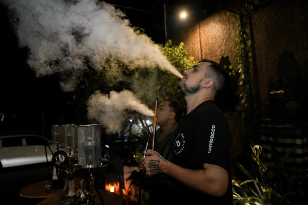 At night outdoors, a person holding a hookah and wearing a black Tombacco shirt is looking up and exhaling a massive, thick white smoke cloud into the air, showcasing impressive smoke production from a shisha session.