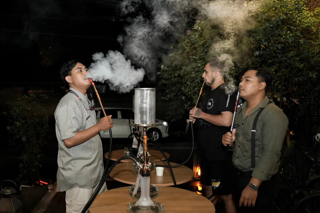 Three friends are standing outdoors at night around several hookahs, sharing a relaxed shisha smoking session together, with one person blowing a satisfying cloud of smoke.