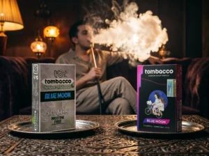 A friendly side-by-side comparison of Tombacco products on a metal tray, helping you decide between herbal or tobacco shisha. It shows a silver box of 'Blue Moon' herbal molasses and a black box of tobacco shisha, while a man enjoys a thick smoke cloud in the background.